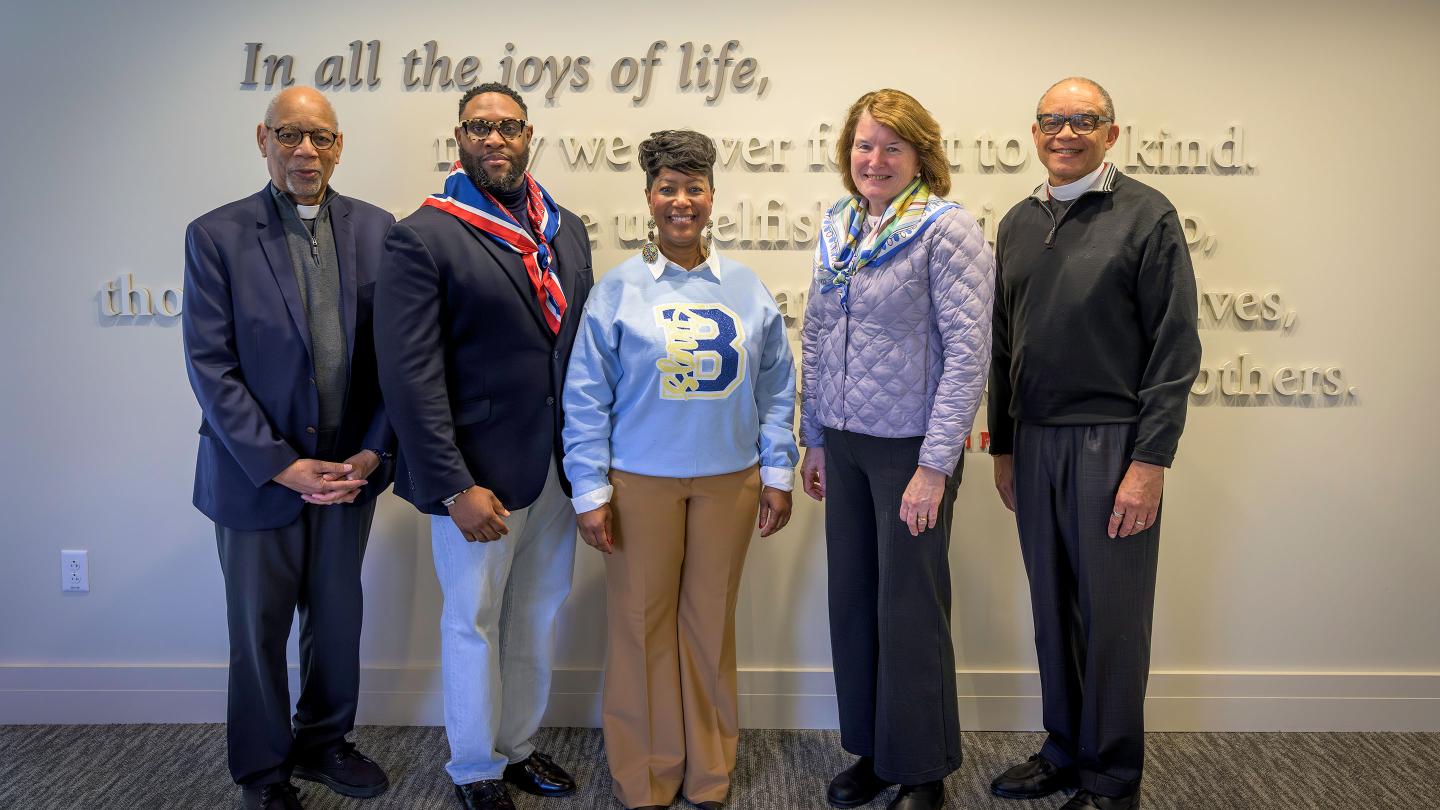 Rector Giles and Rev. Chuck Wynder pose with three visitors from the Bishop Walker School for Boys, standing in front of the School Prayer mounted on a wall in the Fleischner Family Admissions Center.